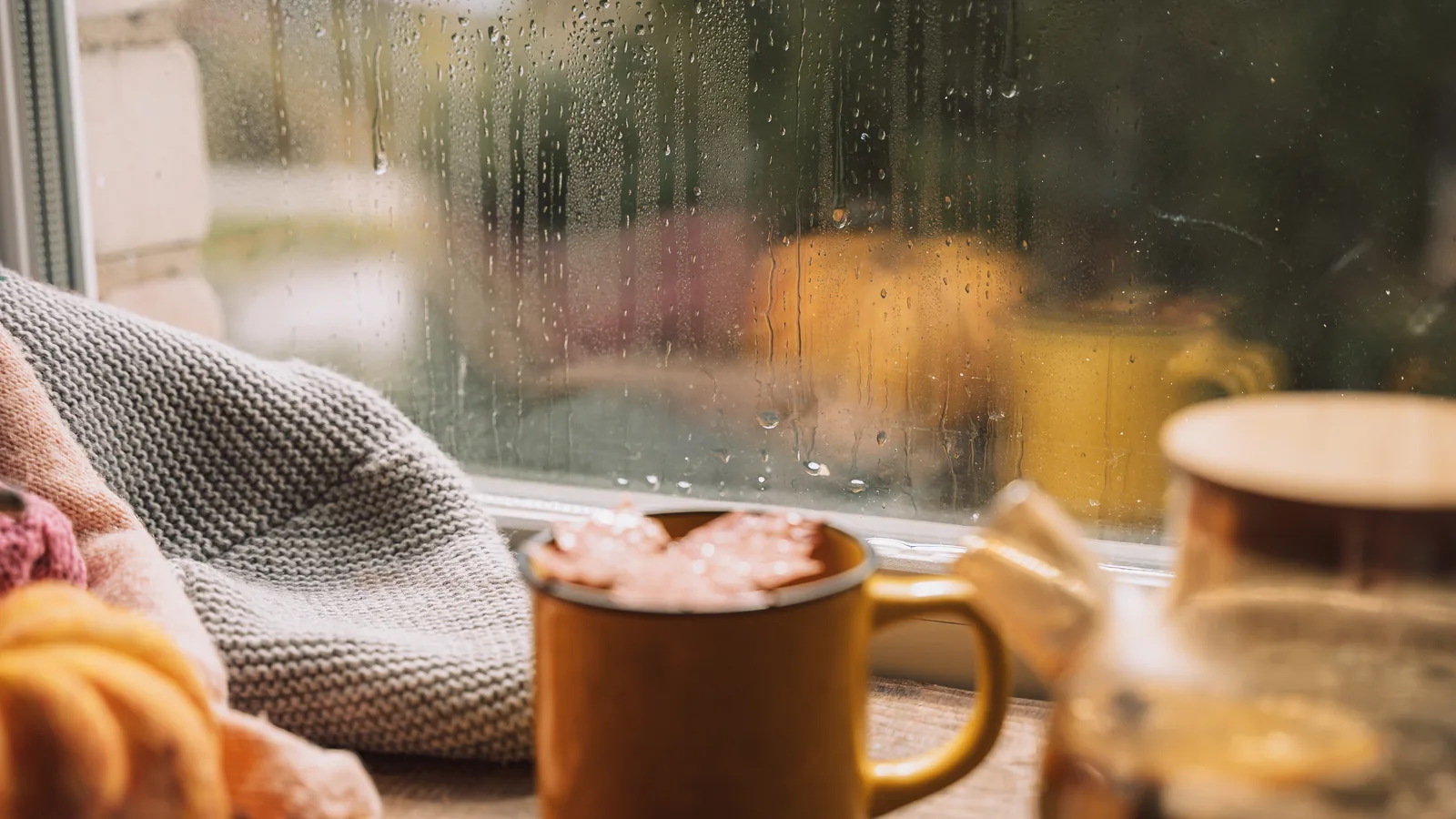 Tasse fumante et plaid en laine devant une fenêtre couverte de gouttes de pluie