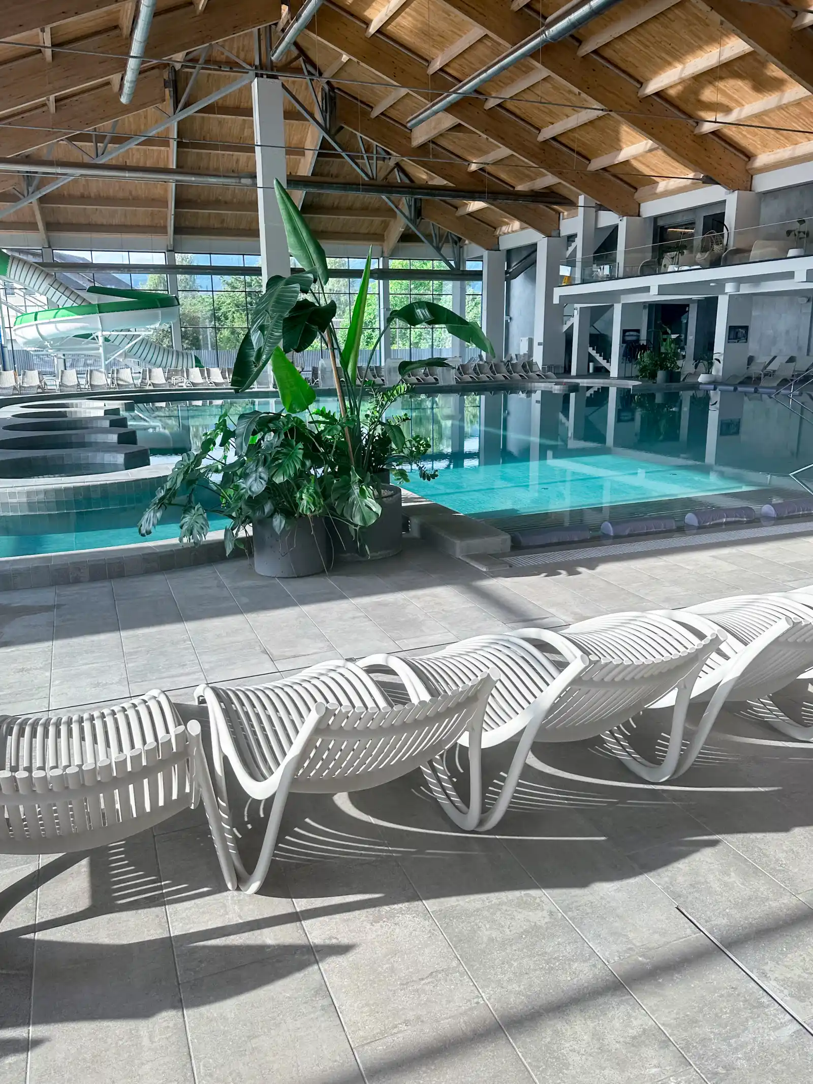 Piscine intérieure sous un plafond en bois, plantes vertes et transats blancs au bord de l'eau