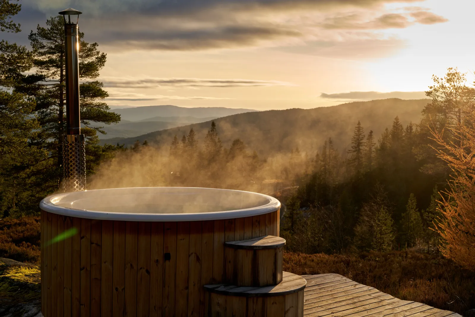 Bain nordique en bois fumant sur une terrasse, face à des collines boisées au soleil couchant